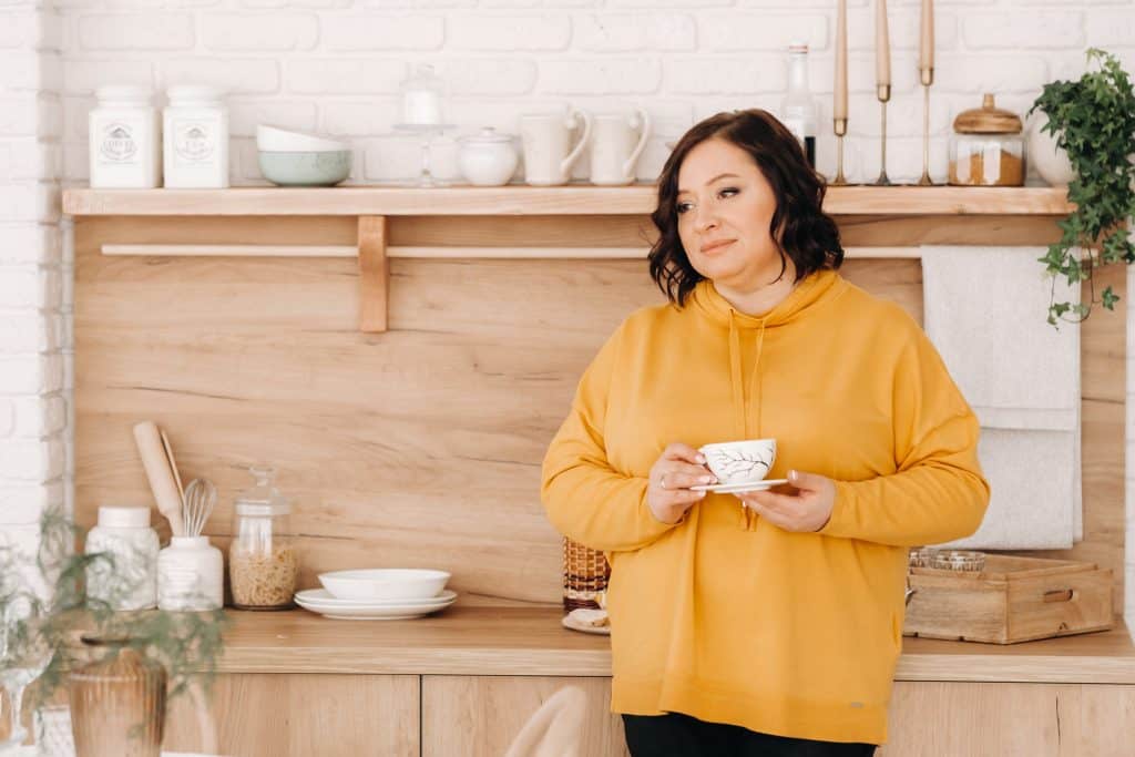 a woman in an orange sweatshirt drinks coffee in the kitchen at home