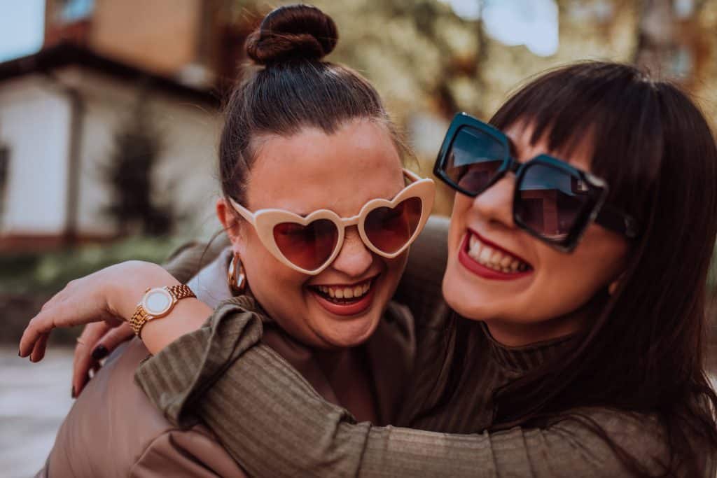 Two women embrace each other emotionally outside on a sunny day while wearing sunglasses.