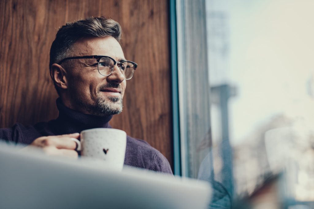 Adult man in sweater sitting in his home and holing a cup of coffee during an online therapy session focused somatic therapy.
