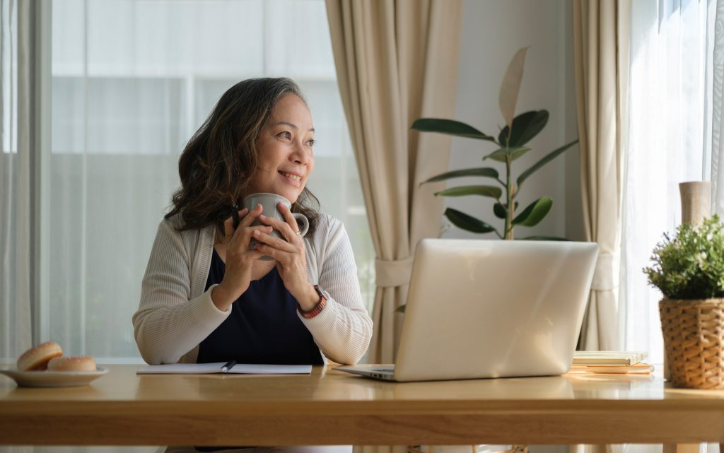 Relaxed middle aged woman holding coffee cup and looking away while waiting to start their online therapy session