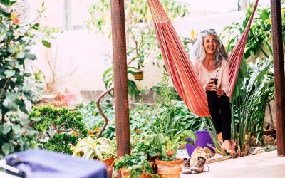 Smiling adult woman with white long hair laughing sit down on an hammock at home in the garden drinking a tea.