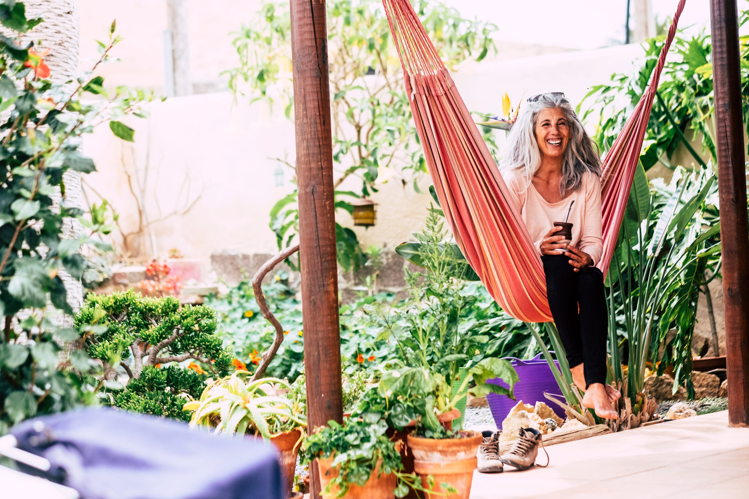 Smiling adult woman with white long hair laughing sit down on an hammock at home in the garden drinking a tea.