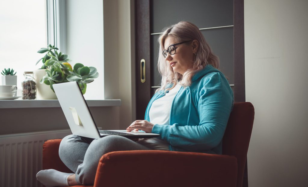 Side view photo of a woman on her laptop while wearing glasses and casual clothes in an armchair getting ready for her online therapy session in Seattle, WA