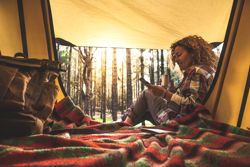 woman using cellular phone sitting outside a tent in the forest with sunlight in background as she engages in self-care