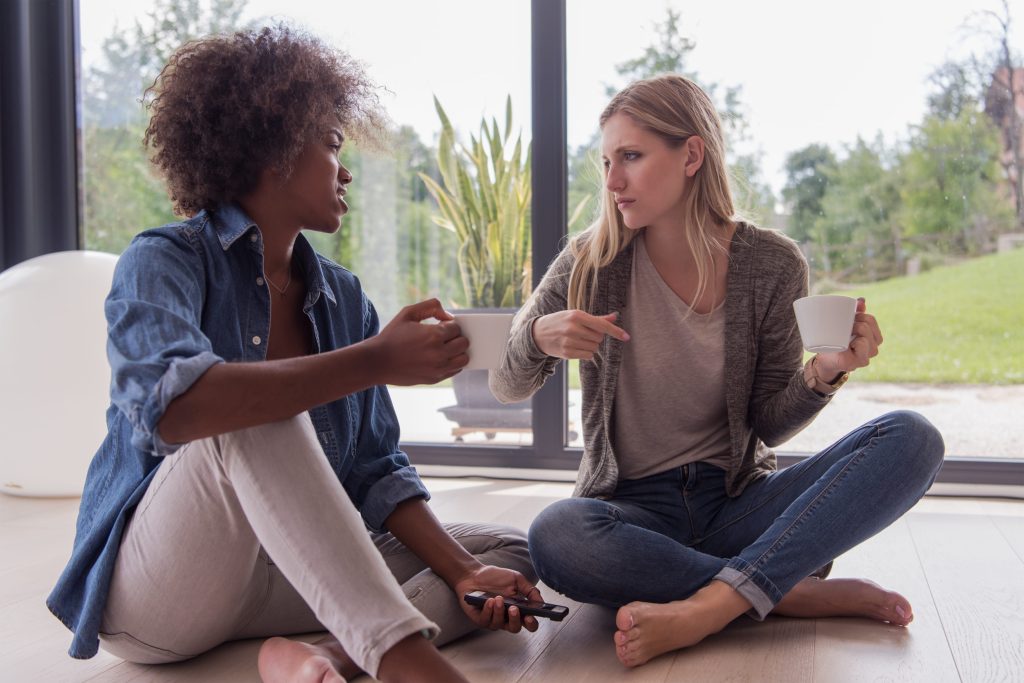 Two women sit on the floor near the window talking while drinking coffee.