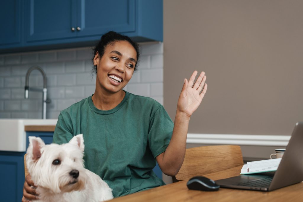 Smiling black woman sitting at the table indoors with her dog on a video call for online therapy via laptop, waving hand